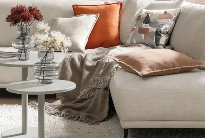 Helles Ecksofa mit beigen und orangefarbenen Kissen, grauer Decke und zwei weissen runden Beistelltischen mit Glasvasen und Blumen auf hellem Teppich.